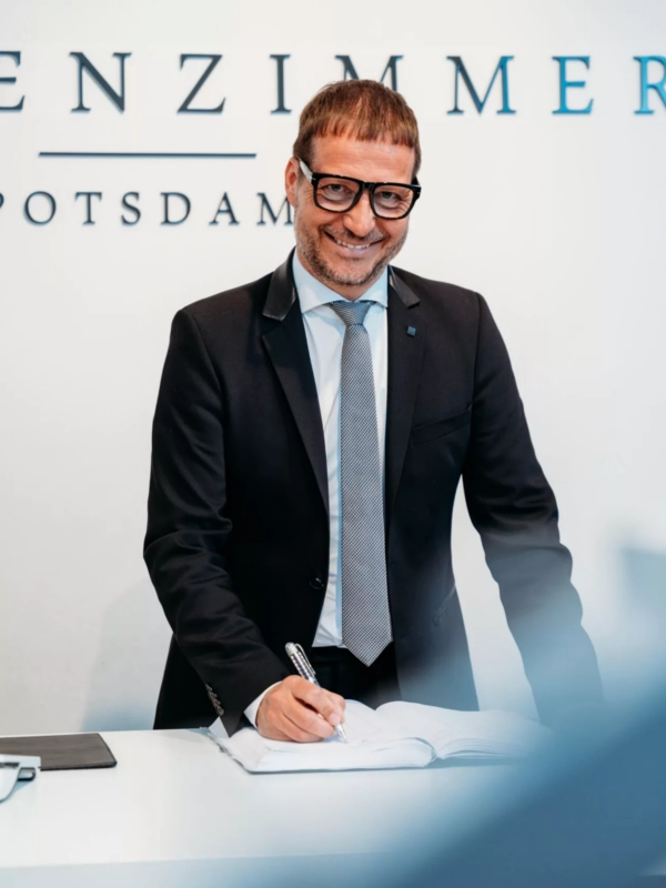 Ein Mann in einem eleganten, modernen Anzug steht lächelnd vor neutralem Hintergrund. Er verkörpert stilvolle Herrenmode und strahlt Selbstbewusstsein sowie Professionalität aus.