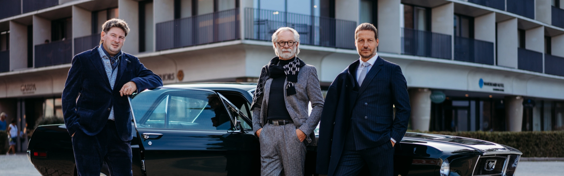 Drei Männer in eleganter Businessmode stehen vor einem schwarzen Oldtimer – sie tragen einen blauen Anzug, einen grauen Anzug im Karomuster und einen dunkelblauen Anzug mit Streifen