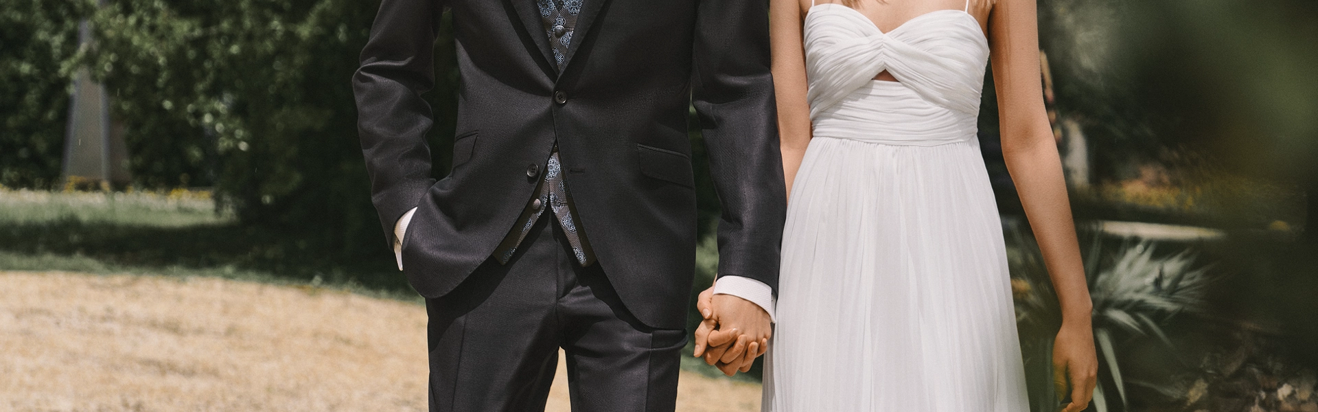 Bräutigam in dunklem Anzug und Braut in weißem, trägerlosem Kleid halten sich im Freien an den Händen. Die Aufnahme vermittelt einen eleganten Hochzeitslook in sommerlicher Kulisse.