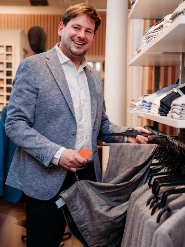Ein lächelnder Mann in einem Herrenausstatter-Laden, der ein Sakko in der Hand hält und von gefalteten Hemden sowie Anzügen umgeben ist. Er trägt eine stilvolle Jacke und vermittelt ein elegantes Flair von Männer-Mode und Stilberatung.